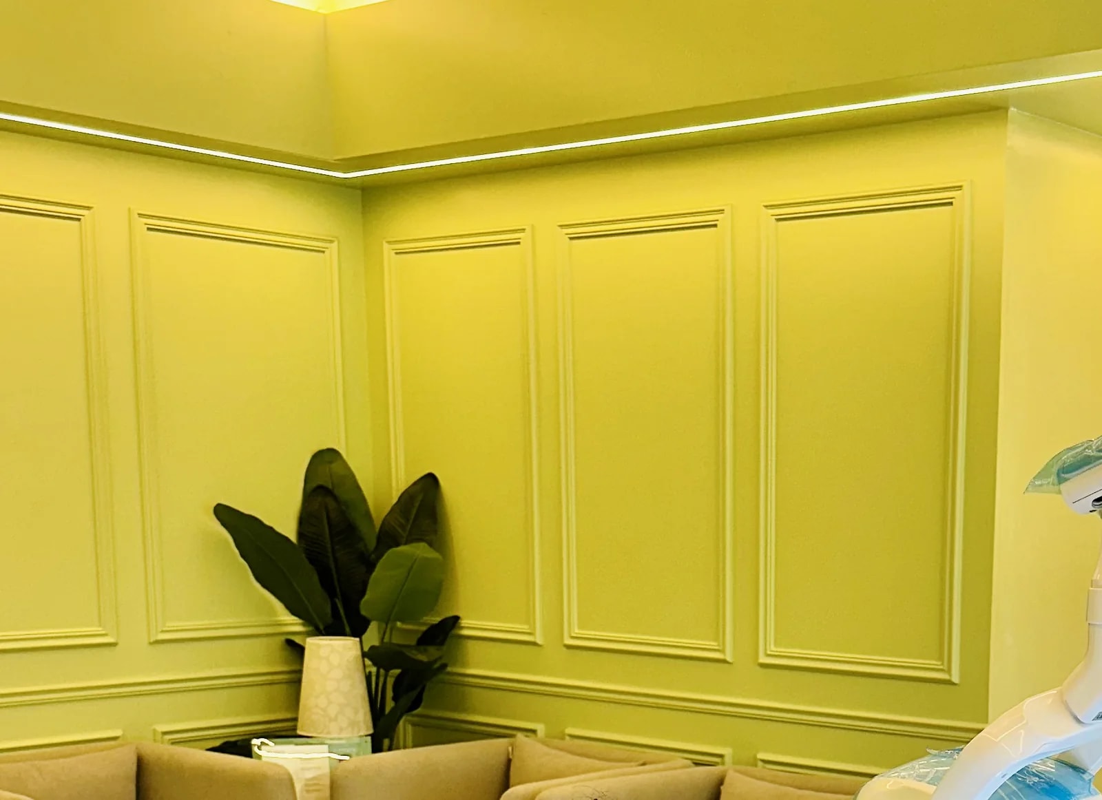 The corner of the treatment room at RH Dental Care Banani: panelled walls, a cove-lit ceiling, a plant and upholstered chairs, with the sheathed arm of the dental light at the right edge.