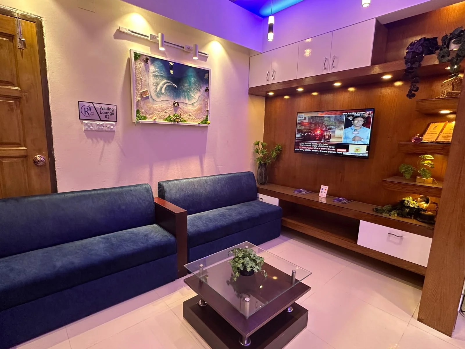 The waiting area at RH Dental Care Banasree: a long sofa, a low table, a wall-mounted screen and lit display shelving.